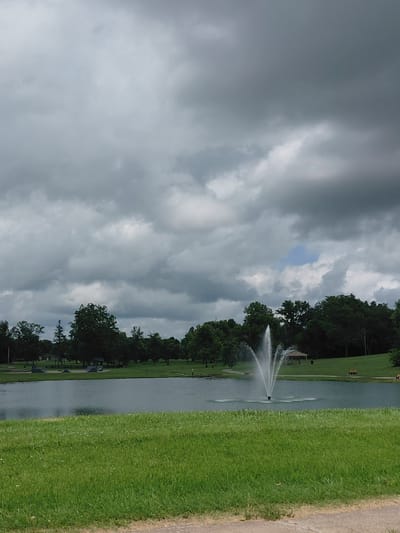 The image depicts a park scene with a pond and a fountain in the foreground. The sky is heavily overcast with dark, dramatic clouds, though a patch of blue sky is visible near the center. A fountain sprays water upwards in the middle of the pond. The pond is surrounded by lush green grass and trees line the background. In the distance, there appears to be a skate park with ramps, a picnic shelter, and park benches. A single, small figure, possibly a person, is visible on the far side of the pond, near the trees. The overall impression is of a tranquil park setting, perhaps on the verge of a storm.