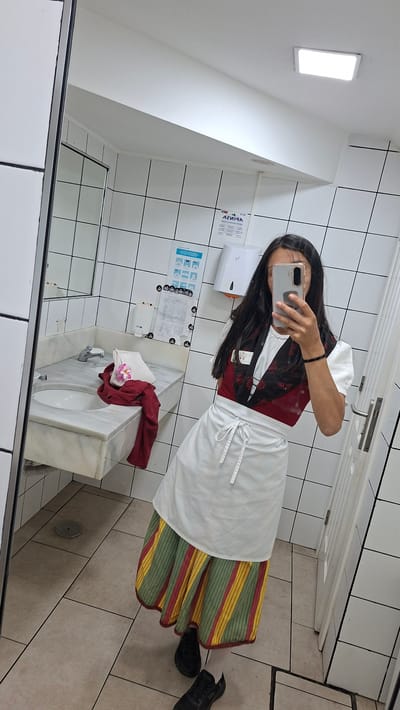 A person is taking a mirror selfie in a tiled bathroom or restroom. The person is wearing what appears to be a traditional or folkloric dress consisting of a white blouse, a red and black patterned bodice, a white apron, and a long, striped skirt with green, yellow, and red stripes. They are holding a smartphone in their left hand, obscuring their face.
In the background, there is a white tiled wall with a hand dryer and what appears to be information posters mounted on it. Below that, there is a white marble sink with a faucet and what appears to be a red garment on it. The floor is covered in light brown tiles. A portion of the reflection shows a door in the background.
Without additional information, pinpointing the exact location within Costa Adeje-playas Del Duque, Spain, is not possible.