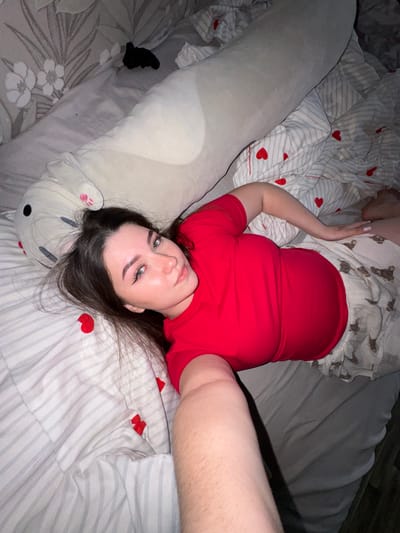 An overhead, close-up shot captures a young woman lying on her back on a bed, taking a selfie. She is positioned towards the left side of the frame, looking directly upwards at the camera with a neutral expression. Her dark hair is splayed around her head on the pillow, and she wears prominent winged eyeliner and light makeup. She is dressed in a plain red t-shirt and light-colored pajamas or shorts patterned with small brown bear figures.

Her left arm is bent at the elbow, with her hand resting near her head, while her right arm is extended upwards, presumably holding the device used to take the photo. A large, light grey plush body pillow, resembling a rabbit or bear with stitched features and a pink paw print on one ear, lies alongside her on the bed, extending from near her head down to her legs.

The bedding consists of a duvet cover with light grey horizontal stripes and scattered red heart motifs. The bottom sheet or fitted sheet visible beneath has a pattern of white flowers on a light grey background. The lighting is dim, suggesting an indoor setting during evening or nighttime. The overall scene conveys a relaxed, personal moment in what appears to be a bedroom in Shirkovo, Russia. There is no visible text.