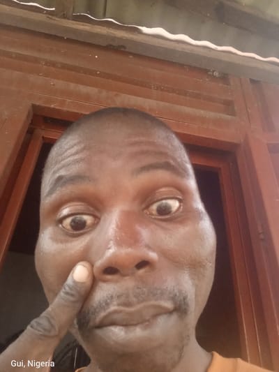This is a close-up, waist-up selfie of a Black man in Kuje, Nigeria. The man has dark skin, a shaved head, and a mustache and goatee. He is looking directly at the camera with his mouth slightly open, and his left index finger is touching his cheek, just below his eye. The background consists of wooden shutters or panels, with what appears to be a portion of a corrugated metal roof visible at the top. The lighting is harsh and casts strong shadows. The man is wearing an orange shirt. The text "Gui, Nigeria" is visible in the bottom left corner of the image.