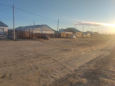 The image depicts a sandy street in the city of Aral, Kazakhstan, at sunset. The sky is a clear blue with a bright sun setting on the right side, casting a warm, golden glow and creating lens flare. Several houses with wooden fences line the street, with one dark-colored car parked in front of a house and a white car further down the road. Power lines and utility poles are visible across the sky. The ground is covered in sand with tire tracks from vehicles. The scene appears to be a residential area in a somewhat rural or developing part of the city.