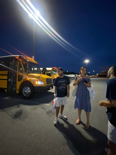 A group of four people are standing in a parking lot at dusk, near a yellow school bus. The sky is a deep blue, with streetlights casting bright beams and lens flares. One person, a man with dark hair, is wearing a black t-shirt with a "Pink Floyd" logo and white shorts, and he appears to be walking towards the viewer. To his right, a woman with dark hair is wearing a blue and white striped off-the-shoulder dress and brown sandals, carrying two white tote bags. Another person, whose back is to the camera, is wearing a dark t-shirt and white shorts, and is standing to the right of the woman. A fourth person, partially visible on the far right, is wearing a dark t-shirt. The school bus is the primary focus of the left side of the image, with its lights on. Another school bus is visible in the background, further back. The ground is asphalt, and shadows are cast by the people and the bus. The scene appears to be set in the city of Halfway, United States of America.
