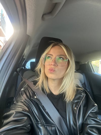 A woman with blonde hair and glasses is in the driver's seat of a car. She is wearing a black leather jacket and a seatbelt. The car is parked, based on the interior view and a glimpse of buildings through the window. It's difficult to pinpoint the exact location in Vallauris, France, but the scene suggests an urban or suburban area given the type of buildings visible. The woman is posing for a photo with her lips puckered.