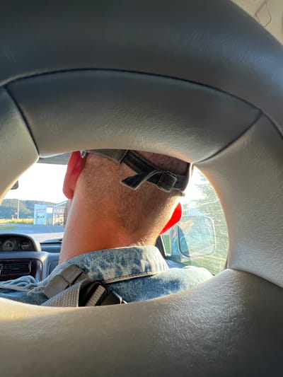 The image, taken from inside a vehicle, shows the back of a person's head and neck, wearing a baseball cap and a denim jacket. The person is seated, and a seatbelt is visible across their shoulder. Through the car window, a building with Cyrillic lettering, "3D РЕГЛАЖ," is visible, along with trees and a hill in the background. The scene is illuminated by sunlight, casting shadows and highlights on the car's interior and the person's clothing. The building appears to be a service station or garage. This scene is located in Dragushinovo, Bulgaria.