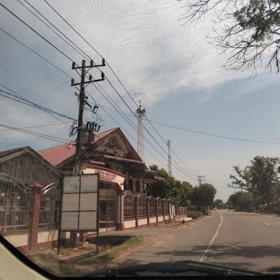 The image depicts a street view from inside a vehicle in Cibrektunong, Indonesia. On the left side of the road stands a building with a red-tiled roof and a decorative facade behind a fence. A sign is erected near the entrance. Behind the building, there is a communication tower, along with utility poles carrying electrical wires. The road extends into the distance, flanked by trees on the right side. The sky is partly cloudy.