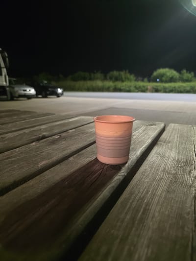 A close-up, low-angle shot of a pink and white disposable cup sits on a weathered wooden picnic table at night. The cup has horizontal ridges around its lower half. The background is dark and blurry, showing a hint of greenery and what appears to be parked vehicles. This scene is situated in Lauenau, Germany.