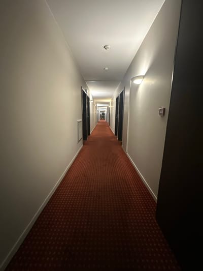 An illuminated hallway extends into the distance, creating a diminishing perspective. The left wall is white, punctuated by circular devices and door frames, while the right wall is covered in a patterned, reddish-brown carpet. The ceiling features lights and door frames. The photograph's perspective emphasizes the hallway's length, drawing the eye towards the light at its end.