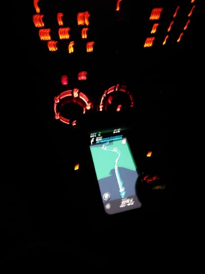 An image of a vehicle dashboard at night. The dashboard features illuminated buttons and dials in red. A smartphone displays a chart, likely showing performance or navigational data, positioned in front of the dashboard. The surrounding environment is dark, highlighting the illuminated elements of the dashboard and phone screen. The photo exhibits motion blur, suggesting the image was taken while the vehicle was moving.