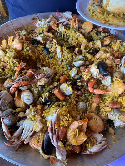 The image shows a large pan of paella, a Spanish rice dish, with various seafood ingredients like shrimp, mussels, clams, and crab, along with sausage and peas. A smaller plate of paella is also visible in the upper left corner. The location is likely San Francisco Lachigoló, Mexico.