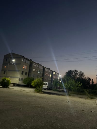 The image depicts a street scene in Şamaldı-Say, Kyrgyzstan, during twilight. A multi-story apartment building dominates the left side of the frame, its windows illuminated with warm light, suggesting occupancy. A streetlamp casts a bright glow on the building's facade. Green trees are scattered along the sidewalk in front of the building, and a white van is parked to the right. The foreground is a dirt road or unpaved area with visible gravel and debris. The sky is a deep, dusky blue, with thin power lines visible against it. There are no people visible in the image.