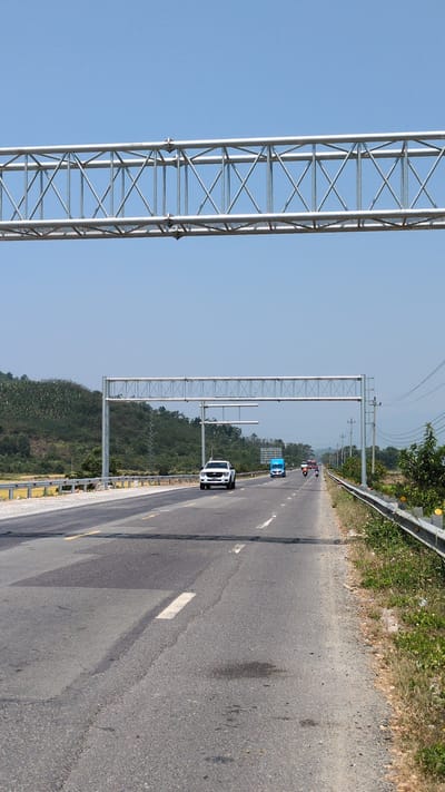 The image shows a section of a highway near Dien Tho, Vietnam. The road is asphalt, with visible wear and tear, and marked with white lane dividers. A white pickup truck is driving in the right lane. Further up the road, a blue car is visible, followed by two motorbikes. Overhead, there are metal gantries spanning the width of the road. The right side of the road is bordered by a metal guardrail and vegetation. The sky is clear. The background shows forested hills.
