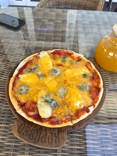 A close-up overhead shot shows a small, round pizza on a wooden cutting board with an inscription. The pizza is topped with melted cheese and red sauce. To the right of the pizza, a glass jar filled with an orange beverage sits on a woven table. A smartphone rests on the table to the left, partially obscured by the pizza. The scene appears to be outdoors, with the woven pattern of the table and wicker chairs suggesting a restaurant or cafe setting. The image is taken in Akhlebaikha (Kovriginskii S/S), Russia.