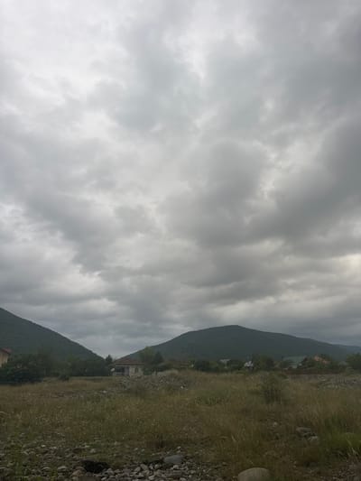 The image depicts a rural landscape with a cloudy sky over Aragvispiri, Georgia. In the foreground, there is dry grass and rocks. The middle ground features a few houses, some with trees and vegetation around them. In the background, rolling green hills and mountains are visible. The sky is overcast with thick, grey clouds. There are no people or animals present in the scene.
