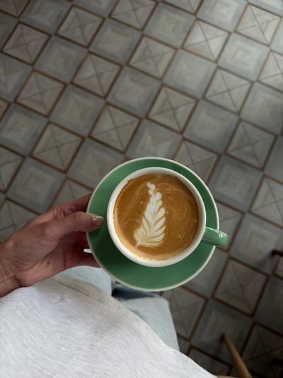 A hand with light brown nail polish holds a green ceramic coffee cup filled with latte art, positioned over a white fabric surface. The coffee cup rests on a matching green saucer. The background is a floor tiled with a geometric pattern of light gray squares outlined in brown. The scene appears to be indoors, possibly in a cafe in Volochysk, Ukraine.