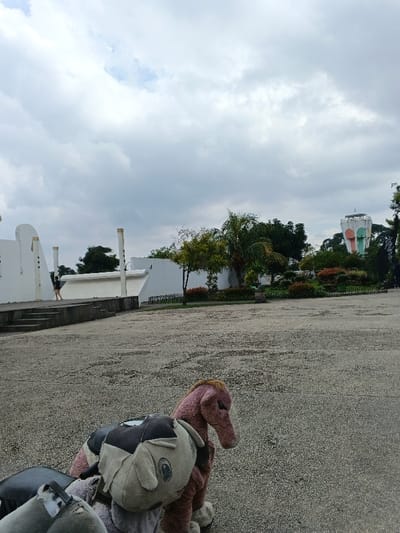 The image depicts an outdoor scene at Văn Miếu Trấn Biên in thành phố Biên Hòa, Vietnam, under an overcast sky. In the foreground, partially visible on a grey, textured paved surface, are several plush toys. The most prominent is a large, light brown or pinkish-brown plush horse, seen from the side facing right. To its left and slightly behind, a grey plush animal head, possibly an elephant or pig, with a dark patch on its head, is visible. Another smaller grey plush object is to the far left.
The midground features an expansive, grey concrete or paved area. To the left, a series of white, modern architectural structures with curved and linear elements, punctuated by several vertical, gold-colored cylindrical posts, rise from a raised platform accessible by steps. A single person, seen from behind in dark shorts and a light top, walks on this raised platform near the white structures. Beyond the paved area, a landscaped garden section is visible, containing various green trees, palm trees, shrubs, and bushes with some reddish-brown foliage. A dark green metal fence or railing separates the paved area from the garden.
In the background, positioned to the far right above the trees, stands a tall, cylindrical water tower. This tower is predominantly white with distinctive vertical sections painted in light green and light orange/pink. The sky above is entirely covered with dense, light grey clouds.