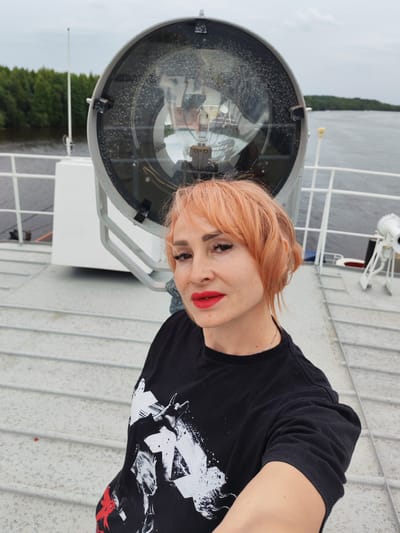 A woman with short, reddish-orange hair and bright red lipstick is taking a selfie on the deck of a ship. She is wearing a black t-shirt with a white graphic print featuring stylized figures and abstract shapes. In the background, a large, industrial searchlight is mounted on a white structure. The ship is on a body of water, with a forested shoreline visible in the distance under an overcast sky. The deck appears to be made of metal with a textured surface. There is a white railing along the edge of the deck. The scene suggests it is daytime, likely overcast given the lighting and sky. The woman's expression is neutral. The location is identified as Novokemskii, Russia.