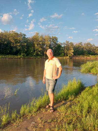 A man stands on the bank of a river, on the right side of the frame, with a forest bordering the opposite bank under a blue sky with white clouds. The man is bald and fair-skinned, wearing a light-colored t-shirt with a graphic on the front, denim shorts, and dark sandals. He has his left hand on his hip and is looking towards the viewer. The river is calm and reflects the sky and trees. Green grass and a dirt path line the riverbank in the foreground. The scene appears to be a natural landscape, possibly a park or a recreational area outside of Chkalov, Russia, given the time of day suggests late afternoon or early evening sunlight.