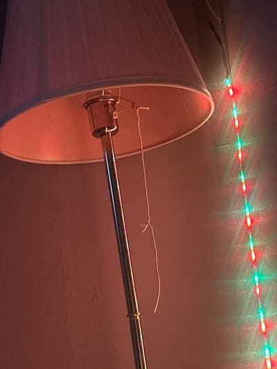 The image shows a lamp with a white shade, suspended by a metal rod, set against a plain wall. A string or cord is attached to the lampshade, extending out towards the right. Above the lamp, a strand of red and green LED lights is affixed to the ceiling. The environment appears to be indoors, possibly a room with warm-toned lighting. There is no information in the image that indicates the location is Kalitinovo, Bulgaria.