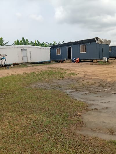 The scene depicts a muddy field in Igrita, Nigeria, with two prefab structures visible. One is white and box-shaped, with a door facing the viewer. The other is dark blue with windows and a door. The ground is waterlogged, and there are patches of green grass. A few chairs and an air conditioning unit are present near the blue structure. Banana trees are visible in the background. The sky is overcast. No people are directly visible.