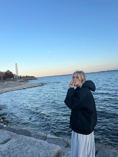 A lone female individual is depicted standing on a rocky shoreline by Lake Ontario in Kingston, Canada, facing partially away from the camera and looking over her left shoulder. She has shoulder-length blonde hair and is dressed in a dark, hooded jacket and light grey sweatpants. Her left hand, adorned with multiple rings, is raised towards her mouth, with lips slightly puckered, suggesting interaction with an unseen small object. In the background, to the left, the prominent tall, white chimney of the Kingston General Hospital power plant is visible beyond a sparse line of trees with autumn foliage. The sky above is clear, transitioning from light blue at the horizon to a deeper blue overhead, with a visible crescent moon. The water of Lake Ontario shows gentle ripples. The scene suggests late afternoon or early evening lighting conditions.