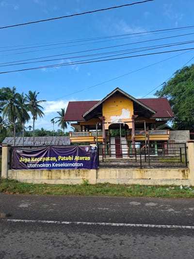 The image depicts a building with a red corrugated metal roof, painted yellow, with some peeling paint, and a partly open structure supported by pillars. A dark blue banner with yellow text is stretched across a fence in front of the building. The banner reads "Jaga Kecepatan, Patuhi Aturan, Utamakan Keselamatan" which translates to "Maintain Speed, Obey Rules, Prioritize Safety." Several palm trees are visible to the left of the building, and dense green trees are on the right. Overhead, several black electrical wires are strung across a bright blue sky with scattered clouds. The foreground shows a paved road with a white line marking. The environment suggests a tropical or sub-tropical location, likely in a rural or semi-rural area, given the presence of palm trees and lush vegetation. The building appears to be an open-air pavilion or a community gathering space, possibly in a state of disrepair or under renovation. There are no people visible in the image. The overall scene suggests a public space with a safety warning displayed.