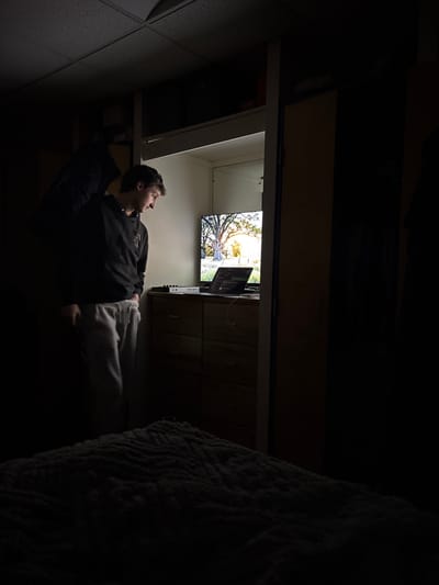 A young man lies on his back in a dimly lit room, gazing up at a flat-screen TV mounted on the wall. The TV displays an image of trees against a bright sky. He's wearing a dark sweatshirt and light-colored sweatpants. The room's environment is dark with a bed and what appears to be a cabinet visible. The lighting concentrates on the TV and the man's face, creating a stark contrast.