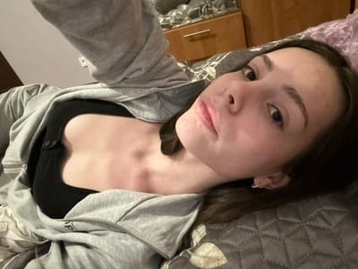 A young woman lies at an angle, facing the camera. She wears a black top and a gray zip-up hoodie. Her dark hair is partially visible against a patterned gray pillow and bedding. Her skin appears fair, and her expression is neutral. A cabinet is partially visible in the lower left. The background also includes a white wall in the lower right corner.