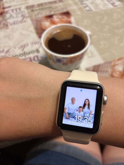 A close-up shot captures a wrist wearing a smartwatch displaying a family photo and the time 19:35. A cup of coffee is visible in the background, on a table with a printed pattern. The image is taken indoors, likely at home, and does not provide any specific information about the location in Zubov, Russia.