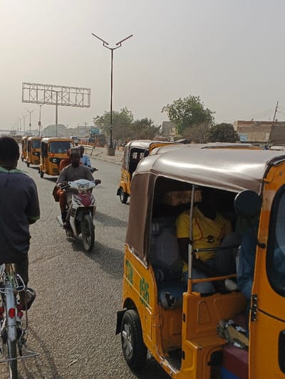 This image captures a street scene in Gobirawa, Nigeria, depicting traffic flow under hazy daylight conditions. In the immediate foreground on the left, the rear wheel and frame of a bicycle are visible, with a person wearing a dark shirt astride it, facing away from the camera. To the right, the front-side of a yellow motorized tricycle (keke Napep) is prominent, featuring a brown canvas roof and showing a male passenger in a yellow shirt seated within.

In the midground, a white and red motorcycle is centrally positioned, ridden by a male wearing a brown garment, with a second person seated directly behind the driver. Behind this motorcycle, a queue of identical yellow tricycles extends down the asphalt road.

The background reveals a hazy, light grey sky. Along the right side of the road, several single-story structures, possibly commercial or residential, are interspersed with green trees. A tall, black street light pole with a dual, angled fixture stands prominently on the left side of the road. Further into the distance on the left, a large, empty metal framework of a billboard structure is visible against the sky. The road surface appears to be asphalt.