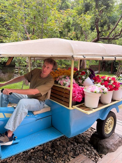 A young man with short brown hair, wearing a green t-shirt and light blue jeans, is seated in the driver's position of a light blue, open-air, three-wheeled vehicle. The vehicle has a beige canopy overhead and is laden with numerous bouquets of artificial flowers in various colors, including pink, yellow, red, and white, displayed in white pots and wooden crates. He is holding onto a black steering handle with his right hand and looking to his left with his mouth slightly ajar. The vehicle is parked on a gravel and stone-covered surface, with wooden planks and large rocks visible in the foreground and background. Lush green trees and foliage form the backdrop. The scene appears to be in an outdoor setting in Mae Rim, Thailand.