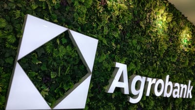 A white illuminated logo, consisting of three interconnected triangular shapes, is mounted on a dense, artificial green wall made of various faux plants and foliage. To the right of the logo, the word "Agrobank" is displayed in large, white, illuminated letters. The wall is textured and appears to be indoors, with artificial lighting highlighting some of the greenery. The image is a close-up view, focusing on the logo and the text. The specific location of this scene in Nurafshan (Toytepa), Uzbekistan is not discernible from the visual information alone.