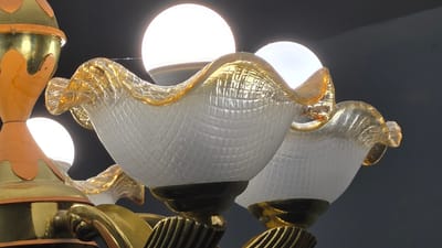 The image is a close-up of a chandelier with multiple lights. The chandelier has ornate gold and orange decorative elements and glass light shades with a textured pattern and gold trim. Three glowing white light bulbs are visible. The background is dark and out of focus. There are no people or actions depicted in the image. The location in the city of Kaka, Turkmenistan is not discernible from the image itself.