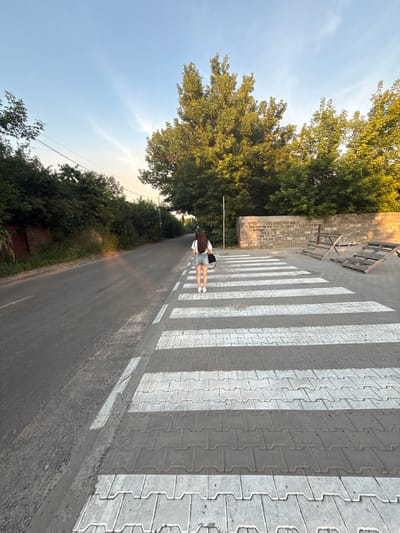 The image shows a person from behind, walking across a pedestrian crossing. The person is wearing a white t-shirt and denim shorts, with a dark bag slung over their shoulder. They have long, dark hair. The pedestrian crossing is made of interlocking pavers with white painted stripes. To the left of the crossing is a road, bordered by lush green trees. Beyond the trees, a brick wall and some wooden structures are visible. The sky is a clear blue, with a few wispy clouds, suggesting it is daytime. The sun appears to be low in the sky, casting long shadows from the pedestrian crossing, indicating it could be late afternoon or early evening. The overall scene depicts a peaceful, possibly rural, environment in Horenychi, Ukraine. No visible text is present in the image.