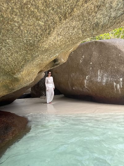 A vertical, medium-shot photograph captures a woman standing in shallow water between large rock formations. The woman, wearing a white dress and sunglasses, is positioned horizontally in the frame. To her left, a large, textured rock formation rises from the ground. To her right, another large rock formation meets the light-colored sand and turquoise water. The scene is set in the city of Lam Kaen, Thailand.