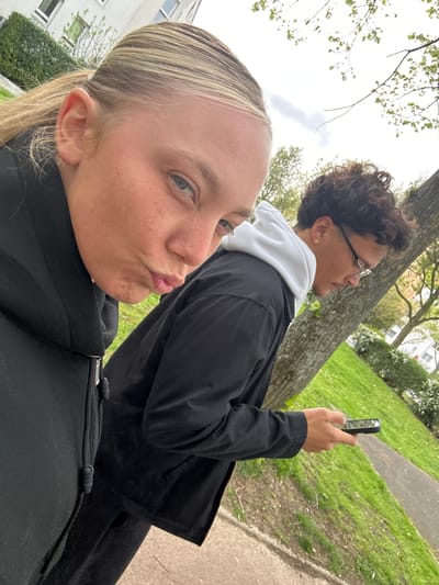 Two individuals are visible in the image. In the foreground, a fair-skinned woman with blonde hair pulled back in a ponytail is making a kissing face at the camera. Behind her is a male individual with curly hair, wearing glasses, a white hoodie, and a dark jacket, holding a cell phone. They appear to be outdoors in a park-like setting with green grass and trees. A building is visible in the background on the left side. The presence of trees lining the sidewalk is consistent with a urban landscape. Given the location specified (Noisiel, France), the buildings and foliage match known characteristics. They could be taking a self-portrait or recording something with the phone.