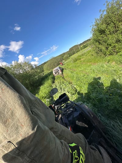 The image depicts a first-person view from a person seated on an ATV in a grassy field near Пепеляева, Russia. The person is wearing olive green pants and a black and neon yellow glove. The ATV is black and muddy. In the background, a white all-terrain vehicle is flipped on its side in a ditch alongside a tree line. The sky is blue with scattered white clouds. There are trees and grass on either side of the field, with power lines visible in the distance.