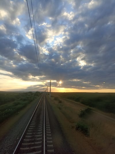 The image captures a dramatic sunset over a rural landscape near Zavoy, Bulgaria. A set of train tracks stretches into the distance, converging at the horizon under a vast sky filled with dark, dramatic clouds interspersed with patches of bright sunlight. The sun itself is a brilliant starburst, peeking through the clouds, casting rays of light across the scene. On either side of the tracks, low green vegetation and fields extend towards the horizon. Overhead, power lines for the train are visible, suspended from poles. The overall impression is one of vastness and the fleeting beauty of nature. There are no people or animals visible in the image.