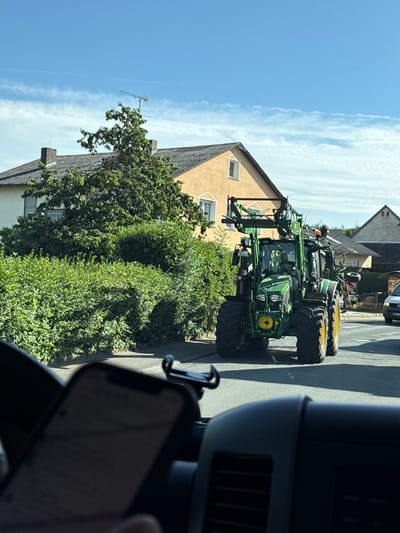 The image depicts a green John Deere tractor with yellow wheels driving down a street in Volpertshausen, Germany. The tractor is equipped with a front-end loader. On the left side of the street, a lush green hedge runs along the property line of a yellow house with a gray tiled roof. A mature tree with dense foliage is situated in front of the house. The sky is blue with scattered white clouds. In the foreground, the interior of a vehicle is visible, with a smartphone mounted on a holder. Another vehicle is partially visible in the background on the right side of the road.