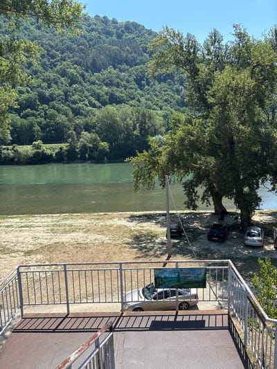 The image depicts a serene riverside landscape on a clear, sunny day. The foreground features a metal staircase leading down from an elevated viewpoint, with a railing overlooking a sandy embankment. Beyond the embankment, a wide, calm river with green, clear water flows, bordered by lush green trees and a forested hillside in the background. Several cars are parked on the sandy area near the riverbank, under the shade of a large tree. A banner with text and a landscape image is attached to the railing in the foreground. The overall scene suggests a recreational area, possibly a park or a picnic spot, situated in a natural setting near Zvornik, Bosnia and Herzegovina.