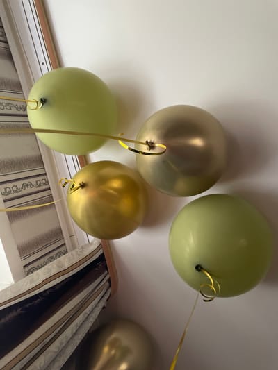 The image shows five balloons, presumably inflated with helium, attached to the ceiling. Two balloons are light green, while three are gold or bronze. Each balloon has a thin gold ribbon hanging from it. The balloons are positioned in front of a window with patterned curtains. The scene appears to be indoors. There is no information to confirm this location is in Ulukulevo, Russia. There is no additional information that is present, such as subjects, objects, or actions.