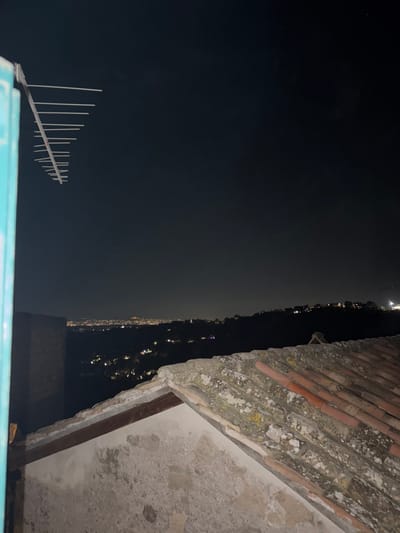 View from a rooftop in Castelnuovo di Porto, Italy at night. The night sky is dark with a few visible stars, an antenna is mounted on a blue-painted structure at the bottom of the frame. In the distance, the city lights are visible in a hazy, orange glow. The rooftop edge is on the right, showing aged bricks and weathered concrete. The bricks are red and covered with lichen and moss. The concrete is stained.