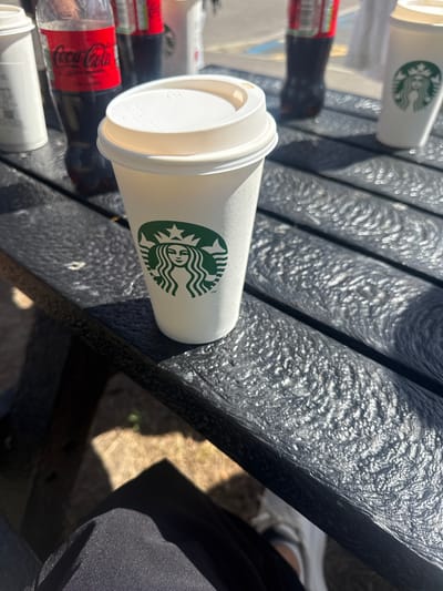 In the city of Hart, United Kingdom, on a dark textured picnic table, a white disposable coffee cup with a white lid sits prominently in the foreground. The cup features the green Starbucks logo. To the left of the coffee cup, slightly out of focus, are two dark-colored plastic bottles, one with a red label that clearly displays the Coca-Cola logo. Further to the right, another white disposable cup, also bearing the Starbucks logo, is visible. The lighting suggests an outdoor setting with sunlight casting shadows and creating specular highlights on the textured surface of the table. The background is blurred, but hints at an outdoor environment with some greenery. The bottom left of the image shows a person's dark clothing and shoes, indicating they are seated at the table.
