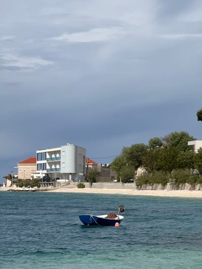 The image shows a small blue boat in a calm, turquoise sea, located in Dugi Rat, Croatia. There is a small, white pebble beach in the foreground, and behind that is a row of buildings and trees. The buildings are a mix of white and red, with some having balconies. The trees are a mix of evergreen and deciduous trees, and they are all green. The sky is a light blue with white clouds. The day is partly cloudy, with a hint of blue sky peaking through. The scene is serene and peaceful.