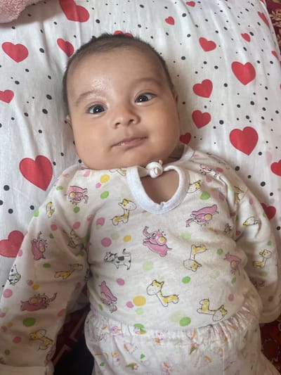 The image shows a baby lying on a white sheet with red hearts and black polka dots. The baby is wearing a white onesie with cartoon animals printed on it, including elephants, giraffes, and cows. The baby's face is visible, with dark eyes and a slight smile. The baby appears to be relaxed and comfortable. The background is slightly blurred, but it appears to be a bed or crib. The lighting suggests that it is daytime. The location is Mashrab, Uzbekistan.