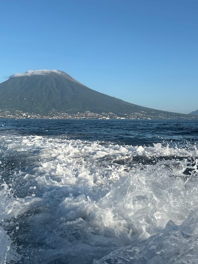 The image depicts a bright, clear day with a large, verdant volcano dominating the background. The volcano's peak is partially obscured by a wispy band of white clouds. Along the base of the volcano, a densely populated coastal town is visible, with buildings of varying sizes and colors dotting the landscape. The town appears to be nestled between the lush green slopes of the volcano and the deep blue waters of the sea.

In the foreground, the turbulent, white-capped wake of a boat is prominently featured, indicating that the photograph was taken from a moving vessel. The water is choppy and filled with spray, suggesting a moderate speed of travel. The sky above is a cloudless, vibrant blue. The overall impression is one of a picturesque tropical setting, likely in the vicinity of Tidore Island, Indonesia, given the distinctive volcanic landscape and coastal settlement.