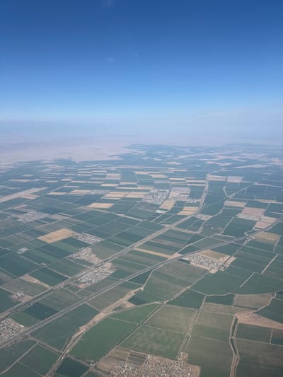 This aerial photograph, captured from an elevated perspective, depicts a vast agricultural landscape dominated by a patchwork of cultivated fields. The fields are organized into numerous rectangular plots, showcasing a variety of crops with distinct shades of green and yellow, suggesting different stages of growth or types of vegetation. Interspersed within the agricultural expanse are small settlements or villages, characterized by clusters of buildings and residential areas. Roads and irrigation channels are visible, indicating infrastructure supporting the agricultural activity and water management. In the distance, a hazy horizon separates the cultivated land from a more arid, mountainous terrain. The sky above is a clear, light blue with a few wispy clouds. The image does not contain any discernible people or animals. While the image itself does not explicitly state the location, if it were to be the city of Shunyi District, China, then this scene would represent a characteristic view of its agricultural outskirts, integrating farming with human settlements.