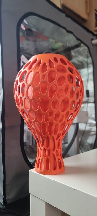 An orange, 3D-printed object with a hollow, porous structure resembling a bell or a mushroom sits on a white surface. The object is illuminated from the front, casting a faint shadow. The background is blurred, showing a dark tent-like structure with a transparent panel revealing the interior. The scene is indoors, likely in a residential or workshop environment, with no indication of an outdoor city setting in Naaldwijk, Netherlands.