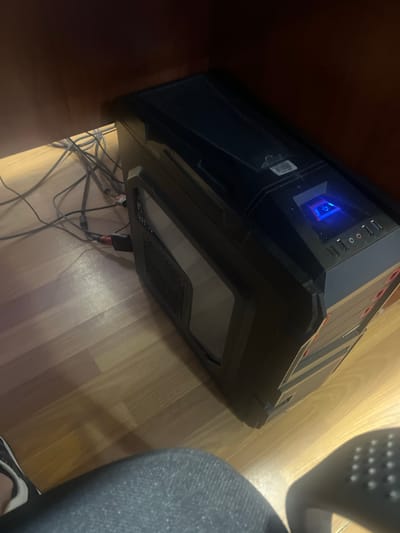 A black computer tower is positioned on a light-colored wooden floor beneath a dark brown wooden desk. Various cables are connected to the computer and are draped across the floor. The computer has a blue illuminated power button on its front panel. A portion of a person's leg, clad in blue denim jeans, and a foot wearing a black and white sneaker are visible in the foreground, suggesting they are seated or standing near the computer. The scene appears to be indoors, possibly in a home or office setting. There is no information in the image to determine the location in the city of Masis, Armenia.