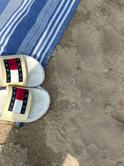 A pair of white Tommy Hilfiger slides rests on a sandy beach, partially overlapping a blue and white striped towel. The slides are positioned side-by-side, with the right slide's logo facing the viewer. The logo features the iconic Tommy Hilfiger flag in red, white, and blue, with "TOMMY" and "JEANS" printed in black text on a white background. A small blue tassel hangs from the right slide. The sand appears slightly damp and textured, with small pebbles and a faint feather visible. The scene evokes a casual, relaxed atmosphere, typical of a beach day. The lighting suggests it is daytime, possibly under sunny conditions given the bright appearance of the sand and slides, though no specific time of day can be determined. The location is identified as Koksijde, Belgium, a coastal town known for its beaches. No specific activity or event is depicted, other than the implied presence of someone resting on the beach.