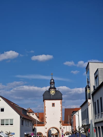 The image depicts the Römertor (Roman Gate) in Obernburg am Main, Germany, under a bright blue sky with scattered white cumulus clouds. The gate, a prominent light-colored stone tower with reddish-brown corner details, stands centrally. It features a dark grey pyramidal roof topped with a belfry-like structure and a golden weather vane. A golden clock face with black Roman numerals is visible on the tower's upper section. An archway at the base of the tower provides passage, through which a red-roofed structure and part of a carousel are visible.
To the left of the tower stands a two-story building with white walls and a reddish-brown tiled roof, featuring multiple rectangular windows, some with blinds. A street sign mentioning "Hauptstraße" is partially visible at its base. To the right of the tower, a modern-style building with a light-colored facade and large reflective windows stands prominently, reflecting the tower's clock face and sky. Further right, another building with narrow, dark vertical windows is partially visible.
The street level in the foreground includes bare, pollarded trees, indicating a specific season. Various street signs are visible, including a circular "No Entry" sign with a white bar and a "3.5m" height restriction sign. Umbrellas or awnings suggest outdoor activity or market stalls. No distinct individuals are clearly visible, but the presence of the carousel and outdoor fixtures implies potential human activity. The scene is illuminated by strong sunlight, suggesting a clear day.